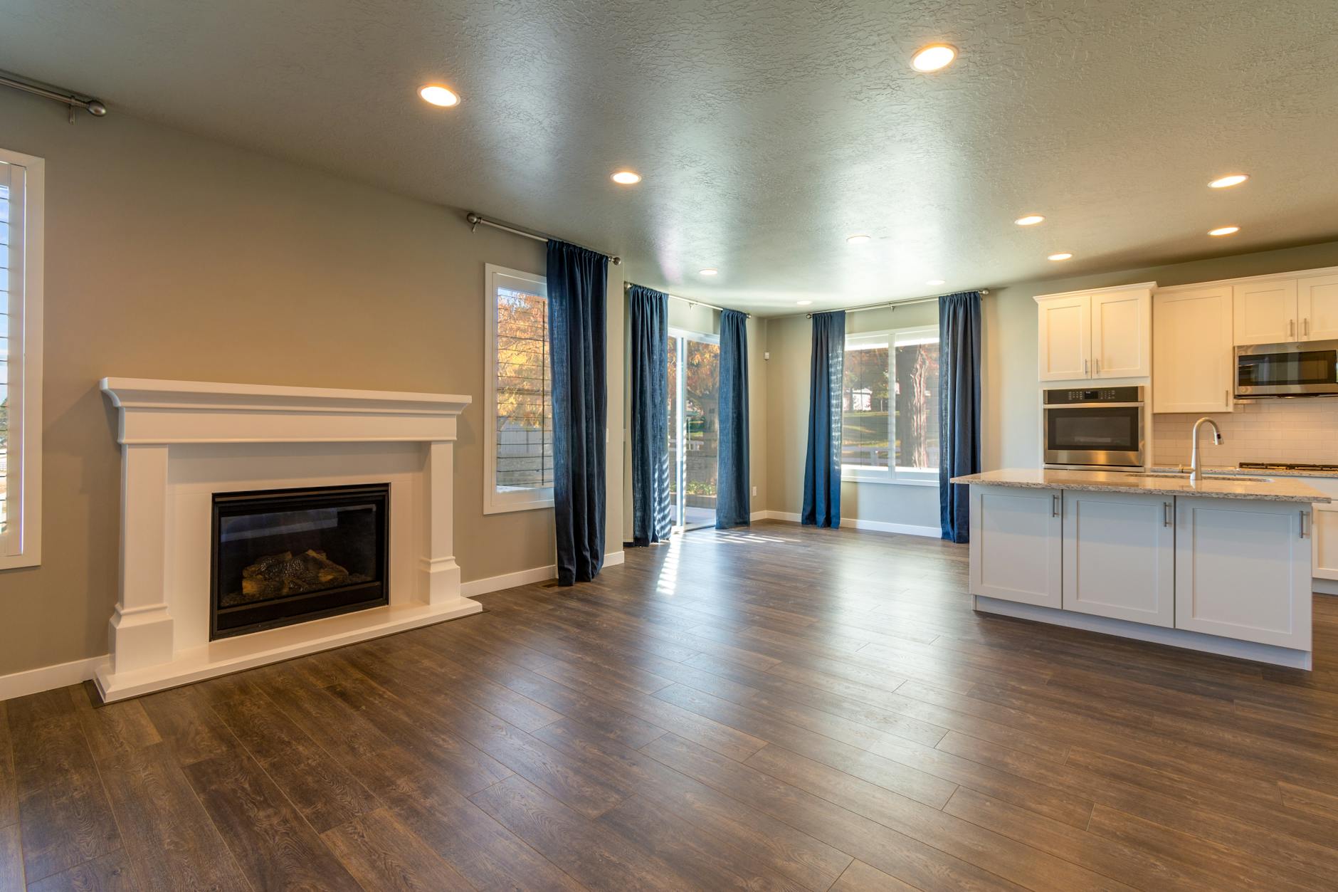 An empty open-plan room with a white fireplace, dark wood floor, blue curtains and a kitchen island