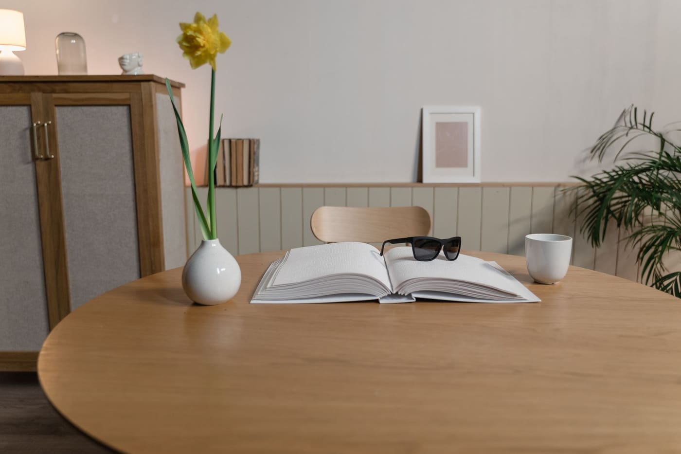 Round wooden table with an open book, a small vase holding a yellow flower and a mug